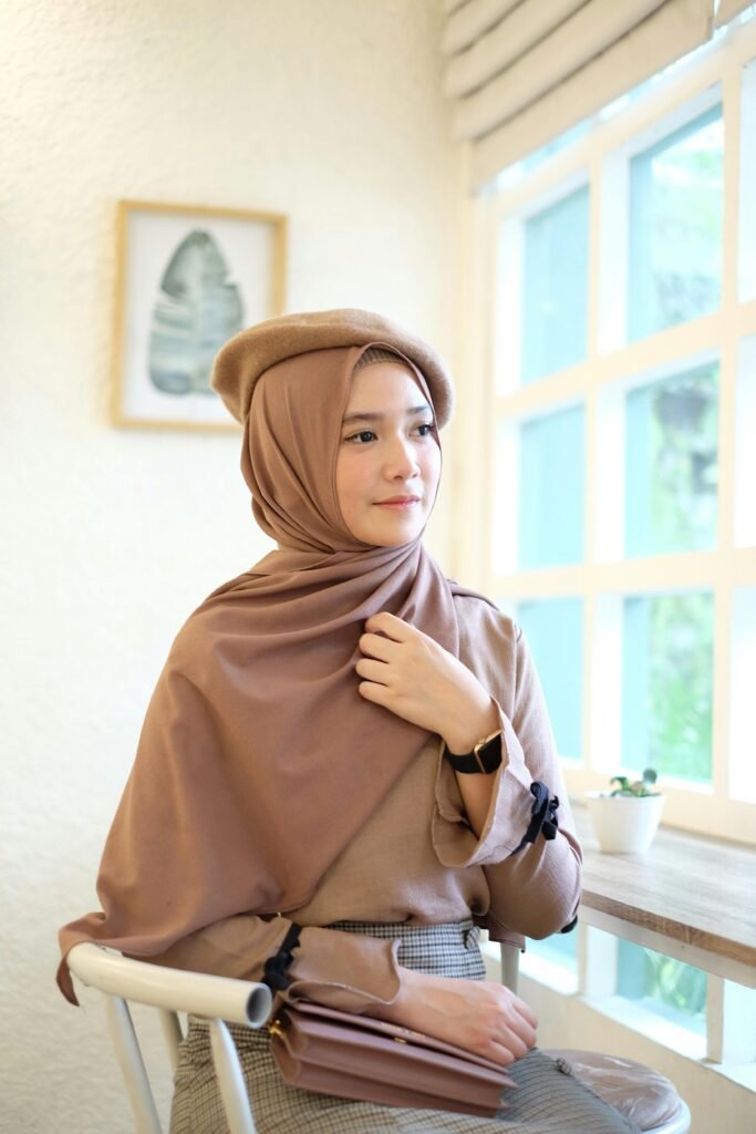 Young woman wearing a hijab and beret, seated by a sunlit window, exuding elegance and poise.